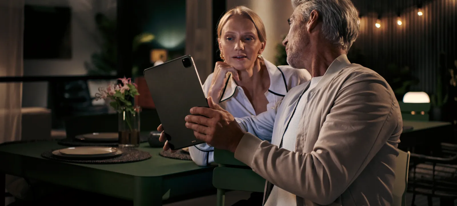 Masada oturan yaşlı çift, tablet tutan kadın, erkek ona bakıyor, dimly lit restoran ayarı.